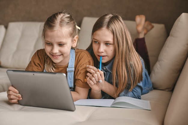 Two beautiful sisters do their homework during quarantine. Children use gadgets for learning. 