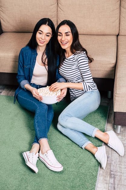 Две красивые улыбающиеся девушки в повседневной одежде едят попкорн и смотрят фильм, сидя дома на диване