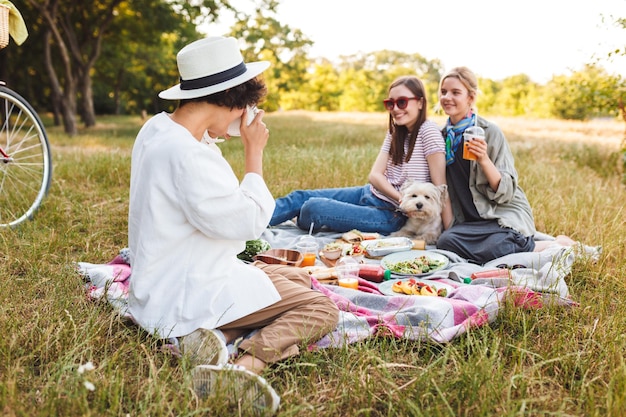 Photo two beautiful smiling girls sitting with little dog on picnic blanket and posing while girl in hat taking photos on camera on picnic in park