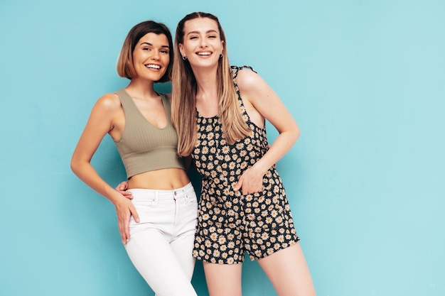 two beautiful smiling hipster female women in summer clothes posing near blue wall in studio