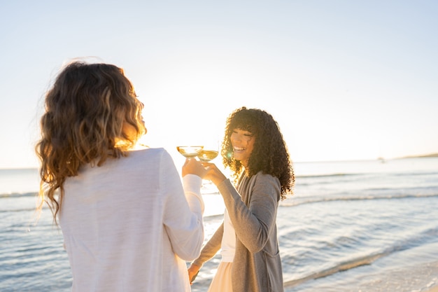 two beautiful trendy young lesbian girlfriends laugh toasting with white wine at sunset 