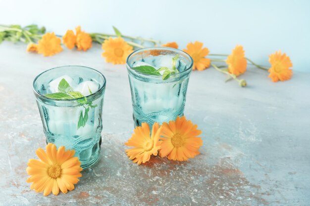 Two beautiful vintage turquoise glasses with cold drink and ice cubes with fresh green mint leaves decorated with marigold flowers on natural colorful stone table Summer concept