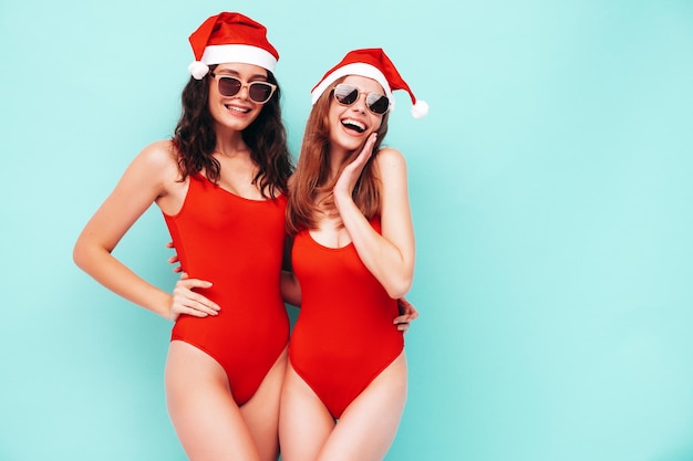 Two beautiful women celebrating New Year.Happy gorgeous female in red sexy body. They having fun at New Year's Eve party.Holiday celebration.Charming models in Santa Claus hats laughing near blue wall