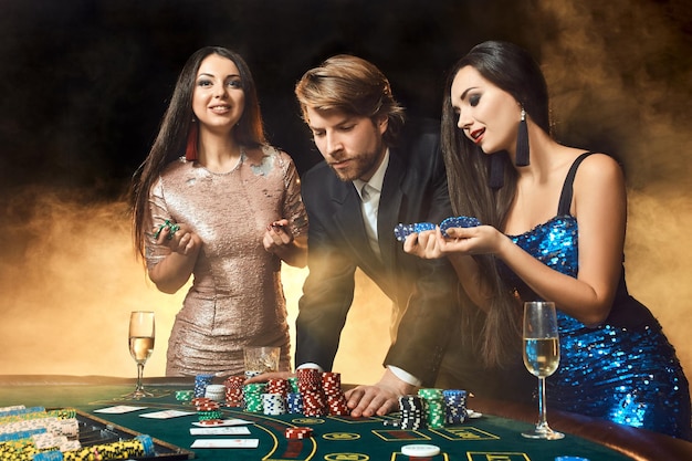 Due belle donne e un giovane giocano al tavolo da poker nel casinò, si concentrano sull'uomo e sulla bruna. passione, carte, fiches, alcol, dadi, gioco d'azzardo, casinò: è intrattenimento. pericoloso gioco di carte divertente per mo