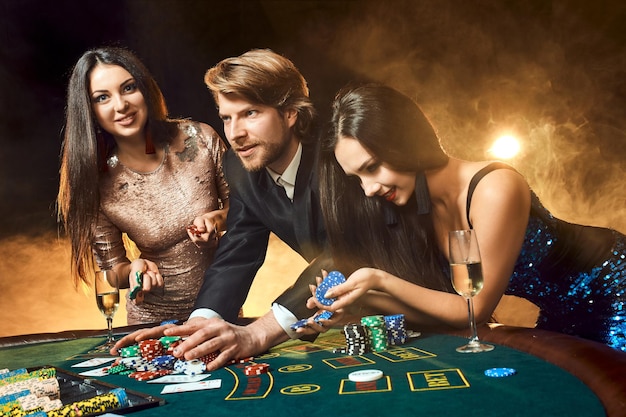 두 명의 아름다운 여성과 젊은 남자가 카지노의 포커 테이블에서 놀고, 남자와 갈색 머리에 초점을 맞춥니다.