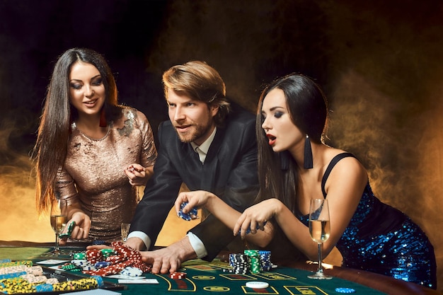 Two beautiful women and young man play on poker table in casino, focus on man and brunette