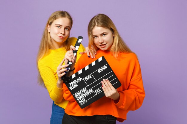 Two beautiful young blonde twins sisters girls in colorful clothes hold classic black film making clapperboard isolated on violet blue wall . People family lifestyle concept. 