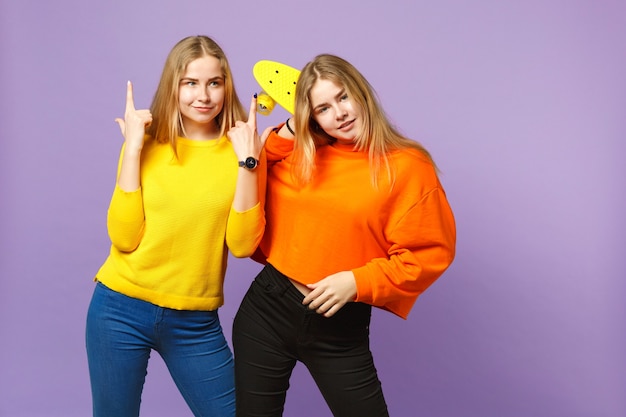 人差し指を上に向けて鮮やかな服を着た2人の美しい若いブロンドの双子の姉妹の女の子は、紫の青い壁に分離された黄色のスケートボードを保持します。人々の家族のライフスタイルの概念。