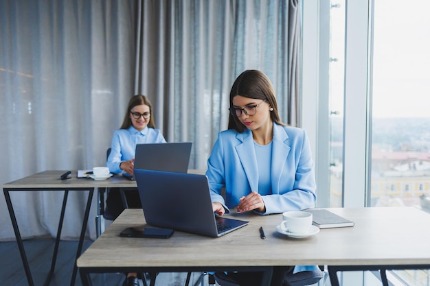 Two beautiful young business women are working and discussing work points in the office Girlfriends at workplaces in the office Large modern office with windows