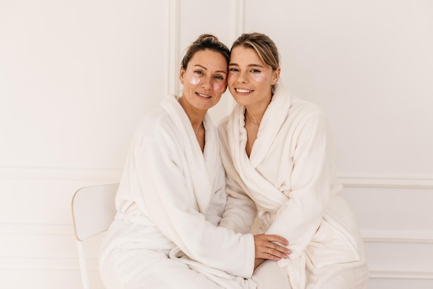 Two beautiful young caucasian ladies take care of skin around eyes with hydrogel patches on white background. Beauty procedure, morning ritual concept