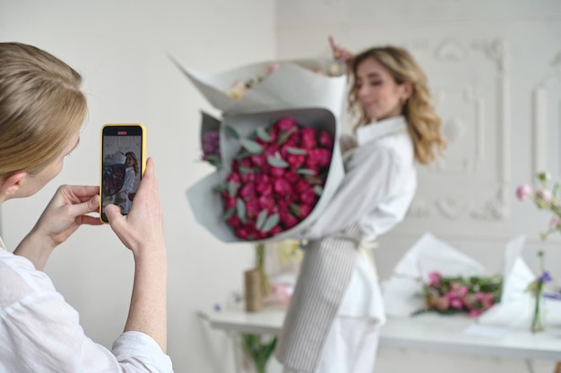 Two beautiful young florists in aprons with bouquets in their hands taking a selfie on a white background