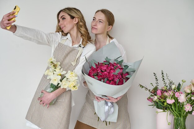 Two beautiful young florists in aprons with bouquets in their hands taking a selfie on a white background