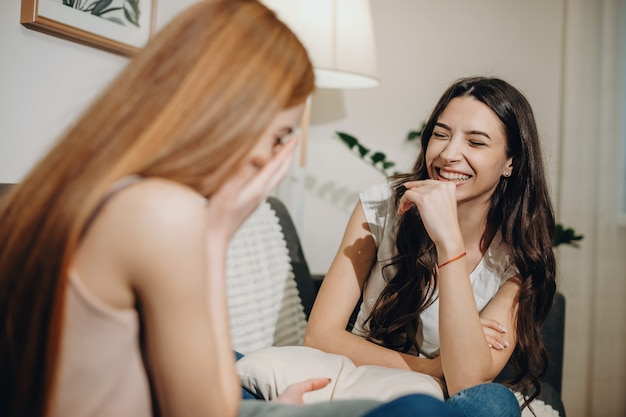 Two beautiful young sister sitting face to face on the couch storytelling while girl with dark hair is laughing with closed eyes.