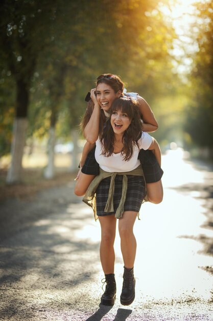 Two beautiful young smiling women with backpacks on their back are making fun during walking on the autumn sunny avenue playing piggyback.