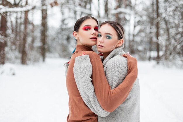 Two beautiful young stylish women model with bright makeup in vintage knitted sweaters hug and warm in a winter park with snow