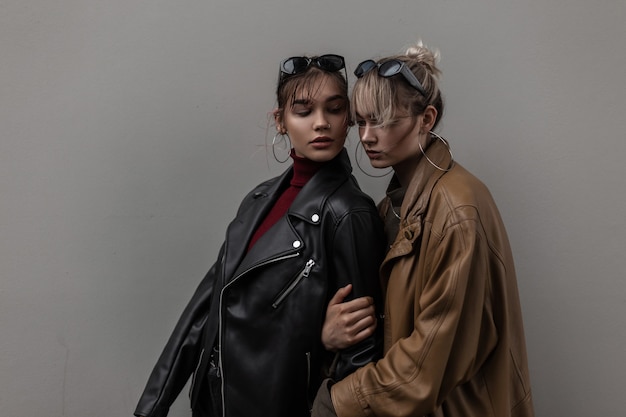 Две красивые молодые стильные женские модели в модных солнцезащитных очках с винтажной кожаной курткой возле серой стены в городе