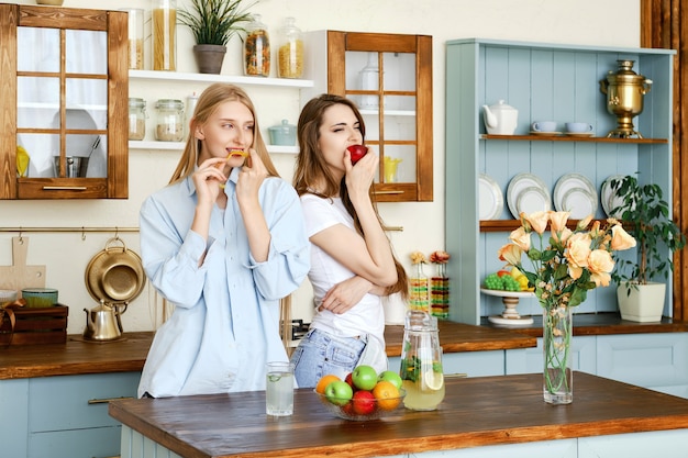 Two beautiful young women eat fruits in the kitchen