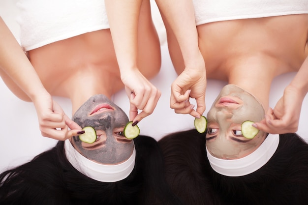 Two beautiful young women holding pieces of cucumber on their eyes and smiling while on the bed