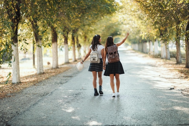 Two beautiful young women with backpacks on their back are walking along the autumn sunny avenue holding hands and looking away.