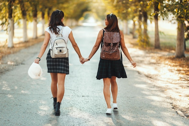 Two beautiful young women with backpacks on their back are walking along the autumn sunny avenue holding hands and looking each other.