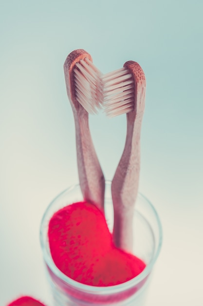 Two beige dental brushes in glass cup with red hearts  on blue white background. Isolated. Toned photo. Love. Valentine day.