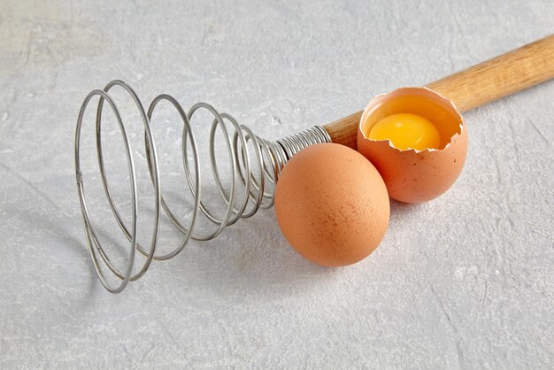 Two beige hens eggs with yolks and a spiral whisk to whip on a gray concrete countertop