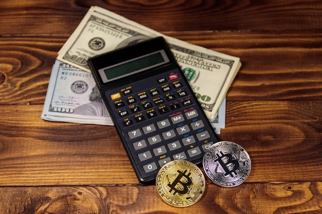 Photo two bitcoins calculator and one hundred dollar bills on wooden desk