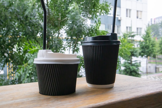 Photo two black paper coffee cups with lids outdoors in summer in sunny weather on a wooden table of a cafe, coffee shop or restaurant.