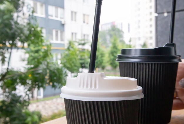 Two black paper coffee cups with lids outdoors in summer in sunny weather on a wooden table of a cafe, coffee shop or restaurant.