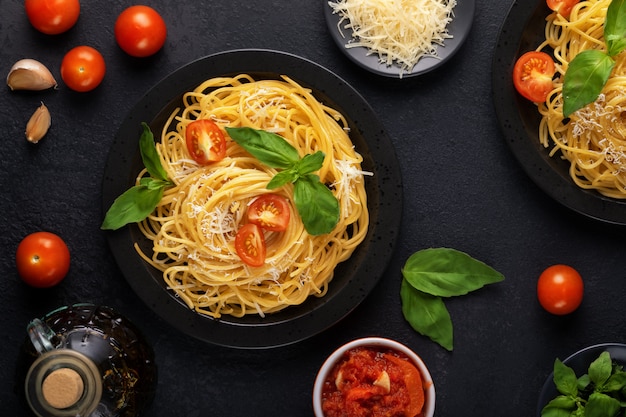 写真 ダークテーブルにベジタリアンの食欲をそそる古典的なイタリアのスパゲッティパスタとバジル、トマトソース、パルメザンチーズ、オリーブオイルの2つの黒いプレート。平面図、水平。