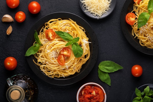 Two black plates with vegetarian appetizing classic Italian spaghetti pasta with basil, tomato sauce, parmesan and olive oil on a dark table. Top view, horizontal.