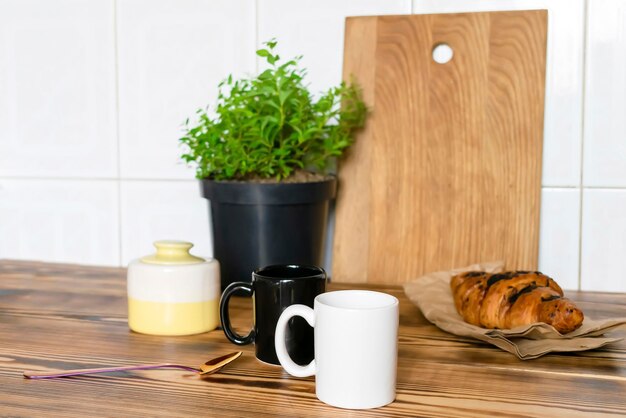 Two black and white espresso coffee cupsfresh baked croissant\
on kitchen table counterutensils dishwarehome green plant in pot on\
wooden shelfearly morning french home breakfastmock up\
template