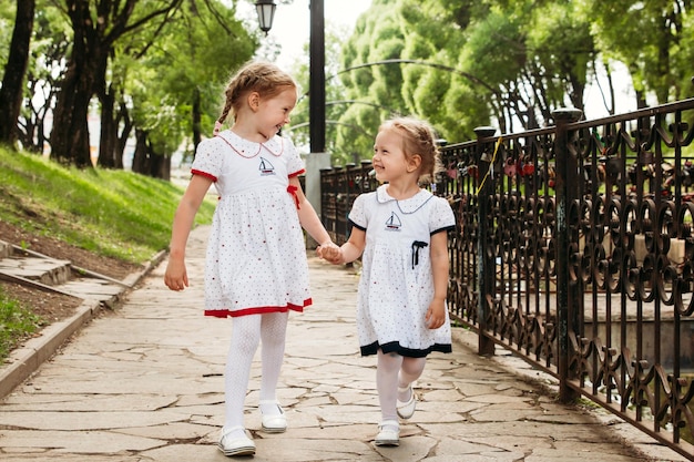 Two blonde Europeanlooking child sisters hold hands and look at each other and smile