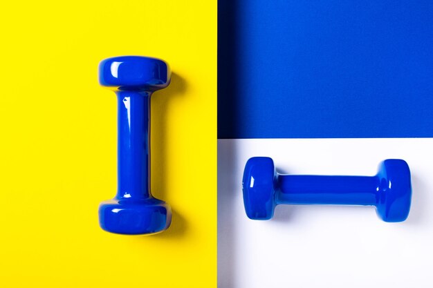 Two blue fitness dumbbells on a white, yellow and blue background. Equipment for home workouts and exercises in the flat lay gym. Sports dumbbell for a healthy lifestyle top view.