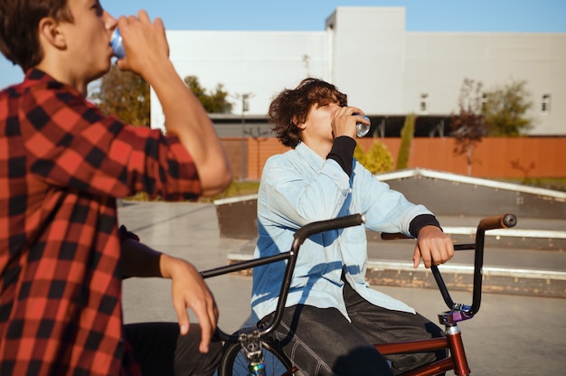 Два байкера BMX отдыхают в скейтпарке после тренировки. Экстремальный велосипедный спорт, опасные велотренировки, уличная езда, езда на велосипеде в летнем парке