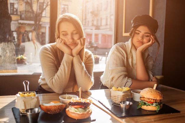 Two bored young women sit at table and look at food. They keep hands under chin. Models are in cafe. Sun is shining outside.