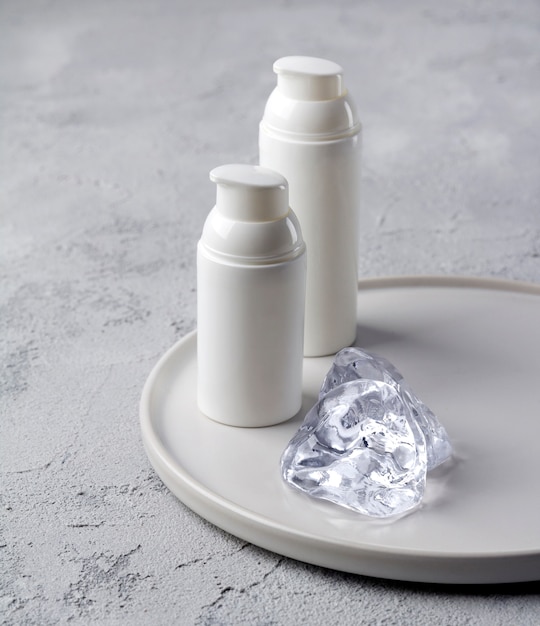 Two bottles of cosmetic cream with ice on gray concrete table.