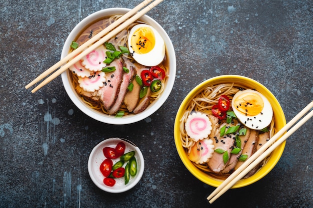 Two bowls of Japanese noodle soup ramen with meat broth, sliced pork, narutomaki, egg with yolk on rustic stone background. Traditional dish of Japan, top view, close-up, concept