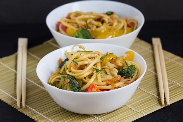 two bowls of stir fry noodles with vegetables and soy sauce on a bamboo mat