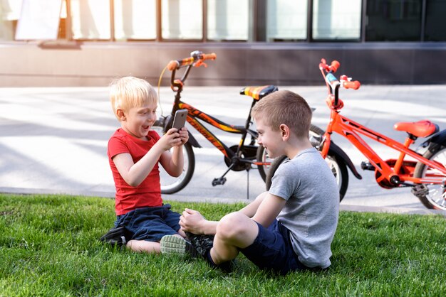 Two boys are photographed on a smartphone while sitting on the grass. Rest after cycling, bicycles in the background