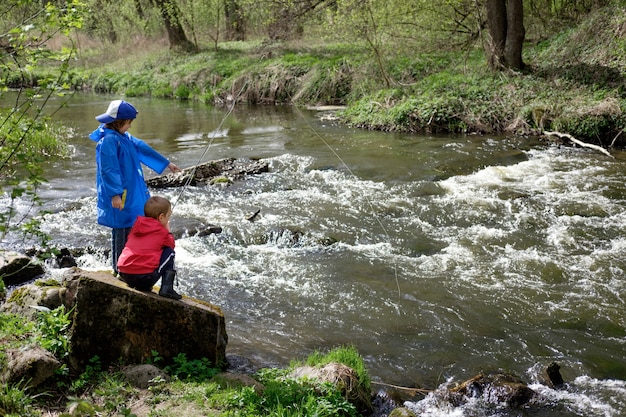 Two boys brothers, dressed in red and blue raincoat, are fishing together on the mount river