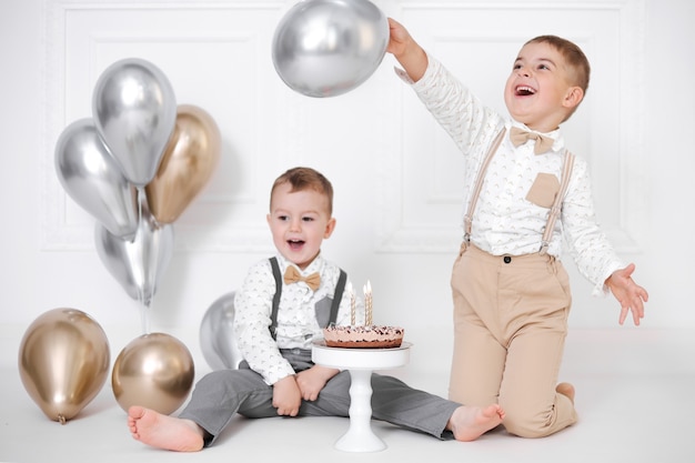 Два мальчика празднуют день рождения, дети празднуют день рождения. Именинный торт со свечами и воздушными шарами. Счастливые дети, праздник, белый минималистский интерьер.