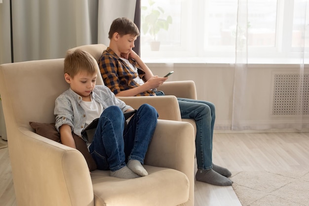 Two boys a child and a teenager sit in beige armchairs in a room during the day play their gadgets