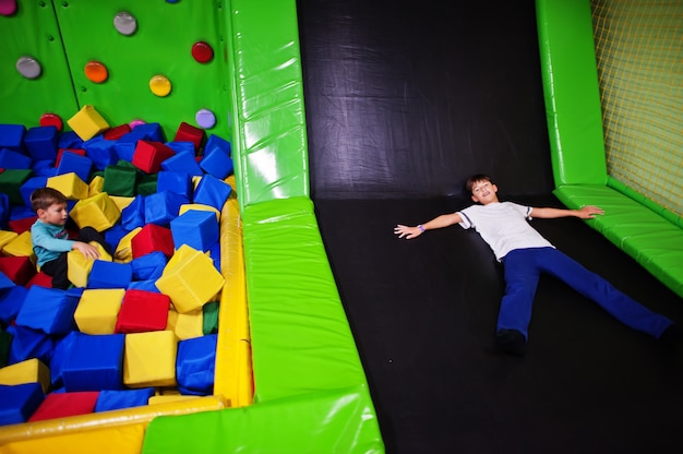 Two brothers lying on pool with colored foam cubes and trampoline in indoor play center.