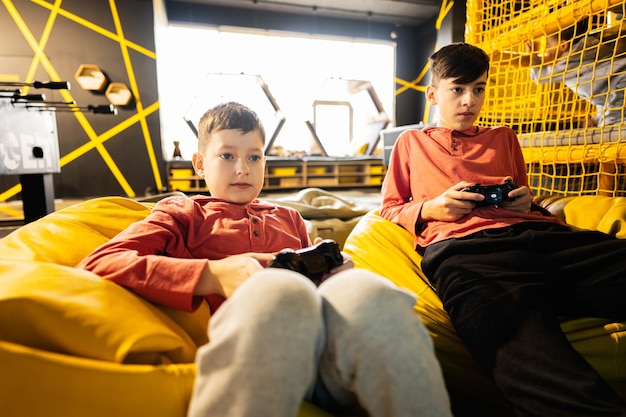 Two brothers playing video game console sitting on yellow pouf in kids play center