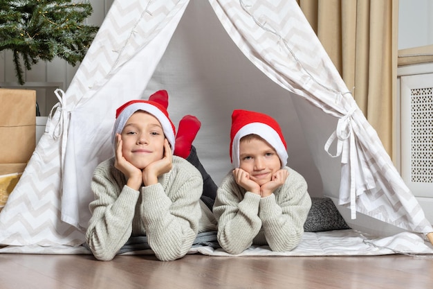 サンタの帽子をかぶった 2 人の兄弟が部屋の子供用テントに横たわり、クリスマスやプレゼントを待ちわびています。