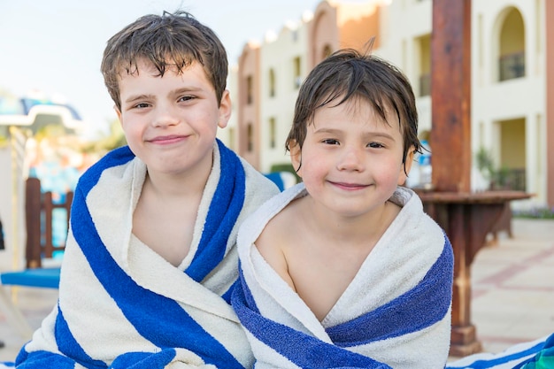 Two brothers wrapped in beach striped towels sitting near swimming pool in luxury resort