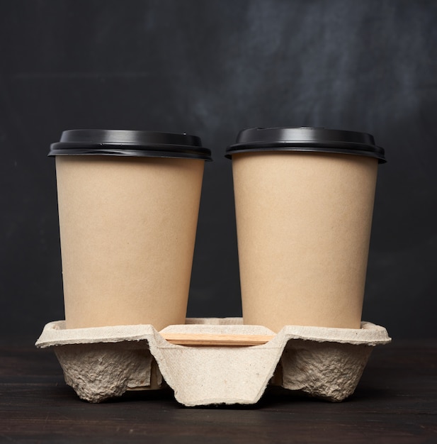 Two brown paper disposable cups with a plastic lid stand in the tray on a wooden table