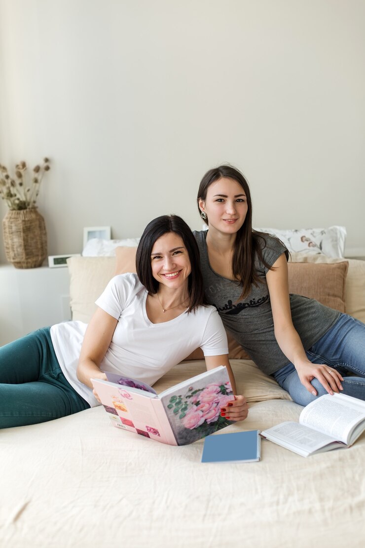 Две брюнетки сидят на кровати с книгами, глядя в кадр.