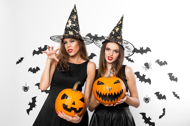 Two brunette girls in black dresses and witch hats hold horror halloween pumpkins on the background with bats .